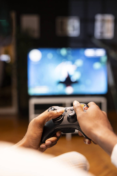Focus shot on man holding controller in apartment, playing videogames on smart TV display in blurry background. Gamer on sofa using joypad to participate in game on console attached to television set - Photo, Image