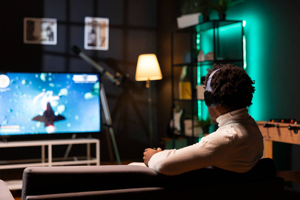 El hombre juega un juego de ciencia ficción para un jugador en la consola de juegos mientras habla con amigos a través de auriculares. Jugador disfrutando de videojuegos con gráficos de alta calidad, utilizando gamepad para navegar por la nave espacial - Foto, imagen