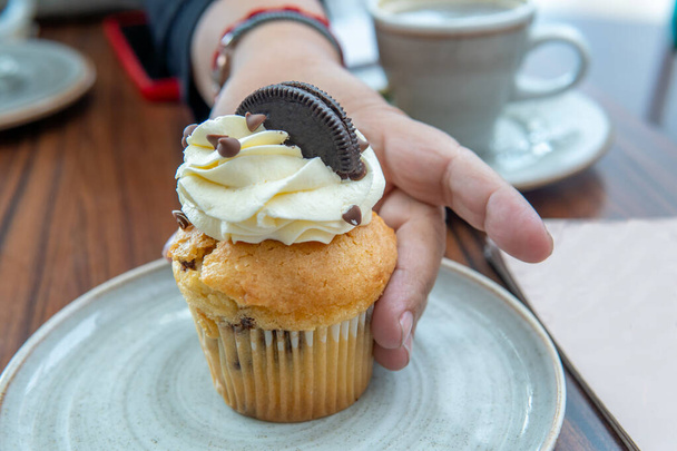gros plan d'une main prenant un cupcake avec crème, gouttes de chocolat et biscuit sur une table - Photo, image