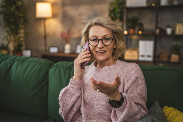 Een volwassen blonde vrouw staan thuis gebruik maken van mobiele telefoon praten - Foto, afbeelding