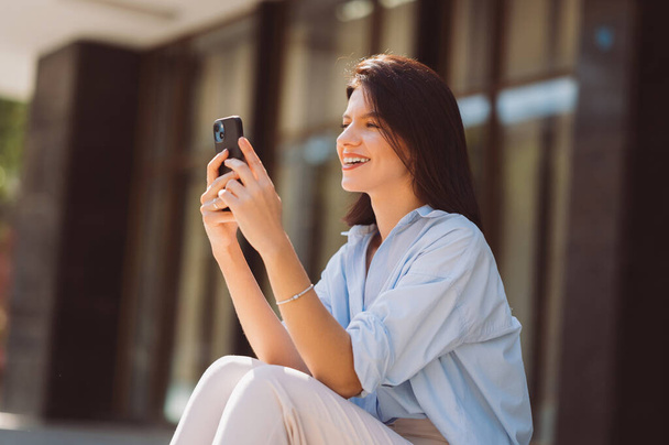 Cheerful young brunette woman is using phone while sitting outdoors on a sunny day. - Photo, Image