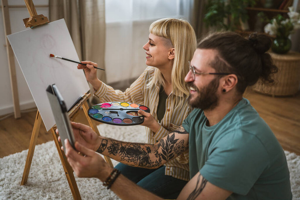 ζευγάρι χρώμα σε καμβά easel στο σπίτι κρατήστε βούρτσα βοηθήσει να μάθετε πώς να το κάνουμε ζωγραφική κρατήσει βούρτσα ευτυχισμένος άνθρωπος και γυναίκα στην αγάπη φίλος και φίλη ή σύζυγος και γυναίκα - Φωτογραφία, εικόνα