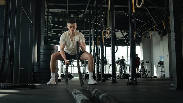Beyazlı fitness adamı spor salonunda halatla çapraz antrenman yapıyor güçlü kaslı sporcu erkek vücut geliştirme sporcusu aktif kardiyo egzersiz dayanıklılığı spor kulübünde vücut geliştirme egzersizi yapıyor. - Fotoğraf, Görsel
