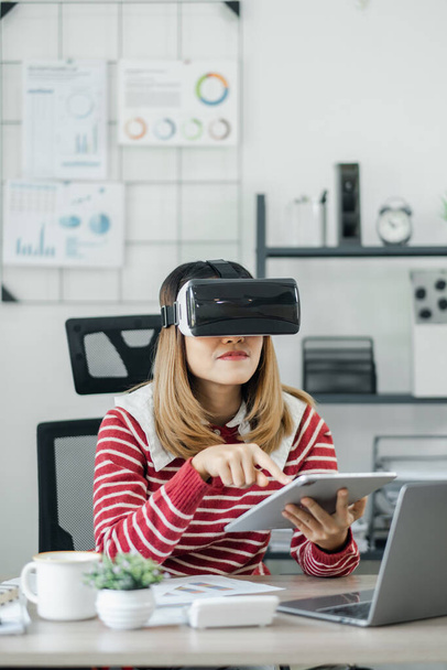 オフィスの設定では,VRヘッドセットを寄付する女性は,タブレットとノートパソコンで同時に作業しています. - 写真・画像