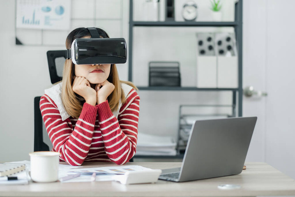 Kobieta siedzi przy biurku z założonymi słuchawkami wirtualnej rzeczywistości, ręce pod podbródkiem, patrząc kontemplacyjnie. - Zdjęcie, obraz