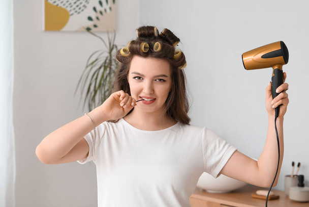 Pretty young woman with hair dryer and curlers in bathroom - Photo, Image