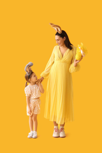 Маленькая девочка с матерью в кроличьих ушах с пасхальным подарком на желтом фоне - Фото, изображение