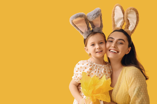 Маленькая девочка с матерью в кроличьих ушах с пасхальным подарком на желтом фоне - Фото, изображение