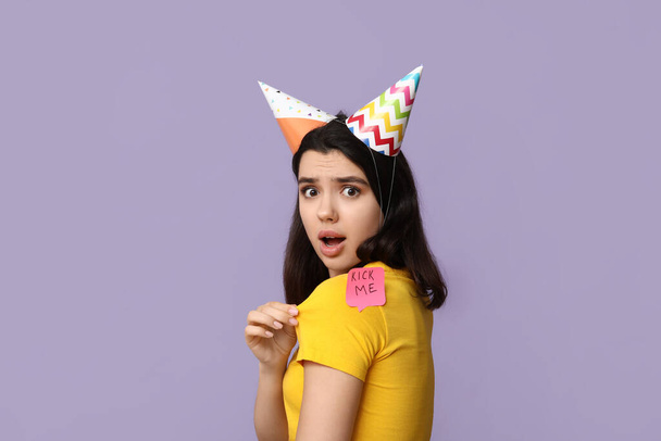 Mooie jonge geschokte vrouw met papieren sticker bevestigd aan haar rug op paarse achtergrond. April dwaze dag viering - Foto, afbeelding