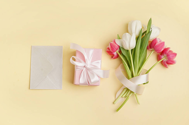 Κουτί δώρου με μπουκέτο από όμορφες τουλίπες και φάκελο σε κίτρινο φόντο - Φωτογραφία, εικόνα