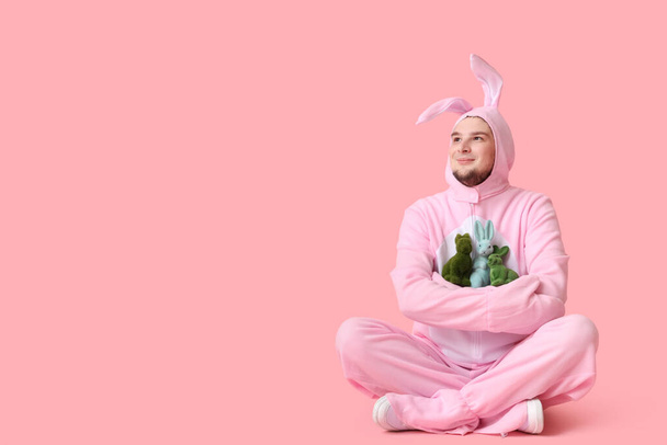 Hombre joven en traje de conejo con conejos de juguete sentado sobre fondo rosa. Celebración Pascua - Foto, imagen