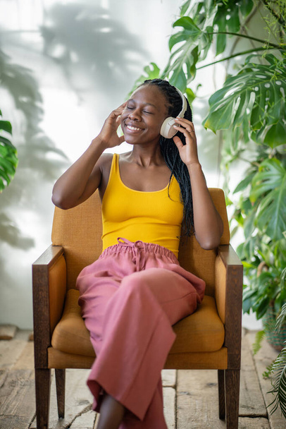 Happy young black woman listen music in headphones, relaxing in armchair in cozy green home with monstera plants, cheerful African American girl spending time, resting at home garden. Enjoy free time. - Photo, Image