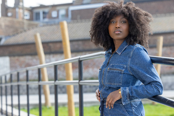 Це зображення зображує стильну афроамериканську жінку з вражаючою зачіскою на афро, впевнено стоїть у міському відкритому середовищі. Шес у джинсовому вбранні, який випромінює випадковий, але шикарний - Фото, зображення