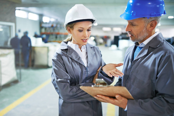 Man, woman and discussion with checklist in factory for quality inspection or safety checks and process monitoring. Team, inventory and clipboard for manufacturing information in printing warehouse - Photo, Image