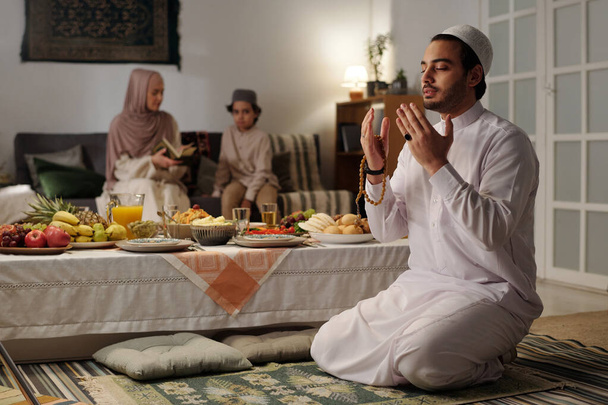Valikoiva tarkennuskuva nuoresta muslimimiehestä istumassa lattialla juhlapöydällä rukoilemassa salahia, hänen vaimonsa ja poikansa lukemassa Koraania taustalla - Valokuva, kuva