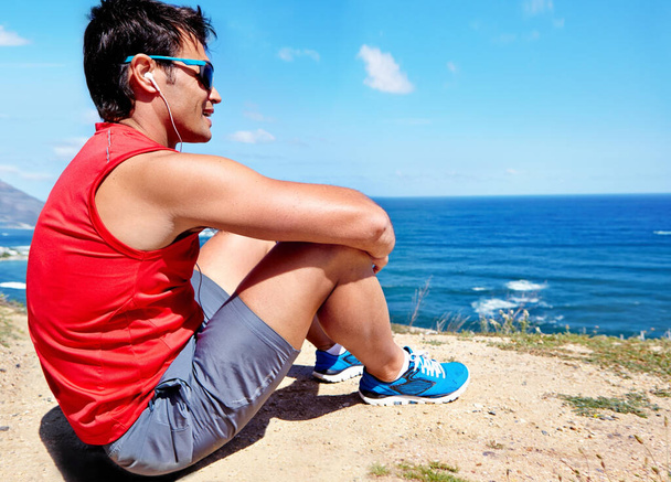 Runner, man and earphones to relax at beach with rest, break or breathing with music in summer sunshine. Person, listening and streaming with subscription with workout, memory and sitting by ocean. - Photo, Image