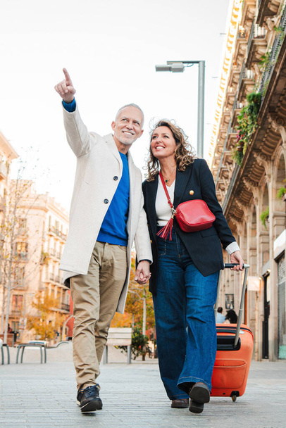 Vertical. Mature tourists pointing and walking with a suitcase on vacations. Middle aged couple having fun sightseeing on a european downtown. Real mid adult married people enjoying a trip on holidays - Photo, Image
