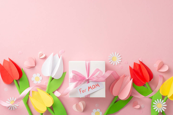 Ремесленное поздравление на День Матери, вид сверху на бумажные тюльпаны, ромашки, подарочную посылку ручной работы с биркой "для мамы", закрепленную лентой, бумажными сердцами и тонкими конфетти на бледно-розовом основании - Фото, изображение
