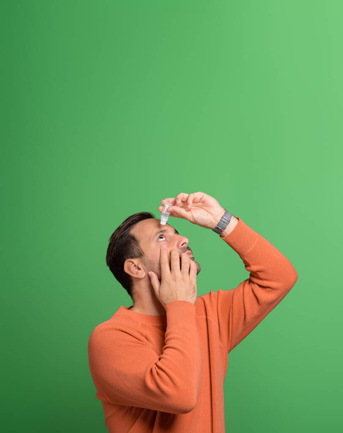 Portrait of overworked young man using eyedropper for treating irritated eyes on green background - Photo, Image