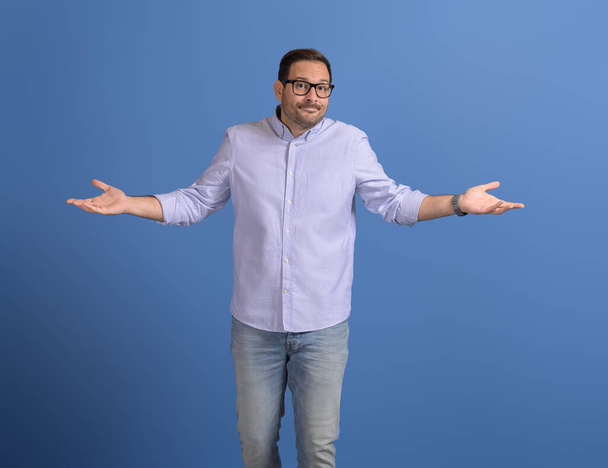 Μπερδεμένος επιχειρηματίας απλώνει τα χέρια και σπρώχνει ώμους ενώ στέκεται πάνω σε μπλε φόντο - Φωτογραφία, εικόνα