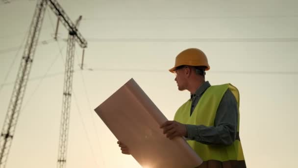 Retrato de cintura hacia arriba del inspector de líneas eléctricas enfocado leyendo dibujos y mirando el pilón de electricidad. Movimiento lento - Metraje, vídeo