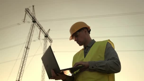 Portret w pasie inspektora linii energetycznej wpisującego na laptopie, stojącego w pobliżu wieży transmisyjnej. Zwolniony ruch - Materiał filmowy, wideo
