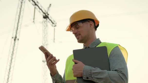 Portrét inspektora elektrického vedení držícího notebook a usmívajícího se při hovoru na mobilu poblíž vysílací věže. Zpomalený pohyb - Záběry, video