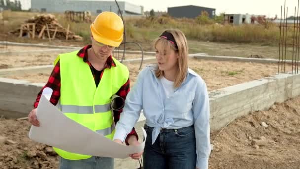 Női mérnök és művezető építészeti tervrajzokat használ, miközben az építés alatt álló új ház alapjait vizsgálja. Lassú mozgás. - Felvétel, videó