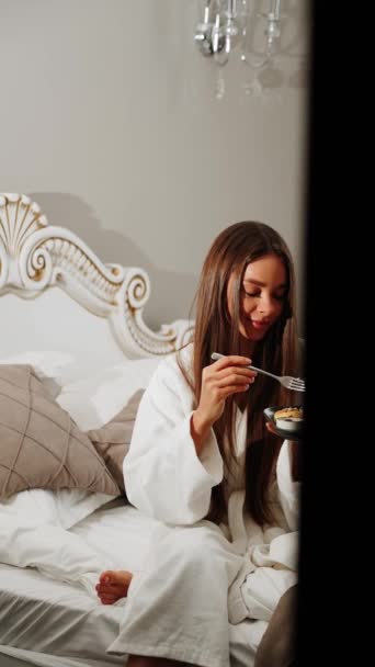 バスローブにいる2人の若い女性がベッドで朝食をとっている. 健康的な朝食チーズケーキ。 ホテルホリデーコンセプト。 バーティカルビデオ - 映像、動画