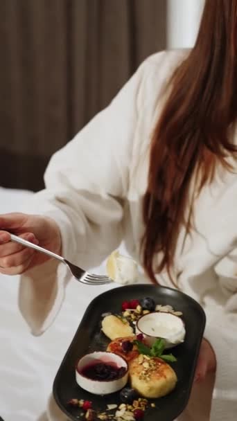 バスローブにいる2人の若い女性がベッドで朝食をとっている. 健康的な朝食チーズケーキ。 ホテルホリデーコンセプト。 バーティカルビデオ - 映像、動画