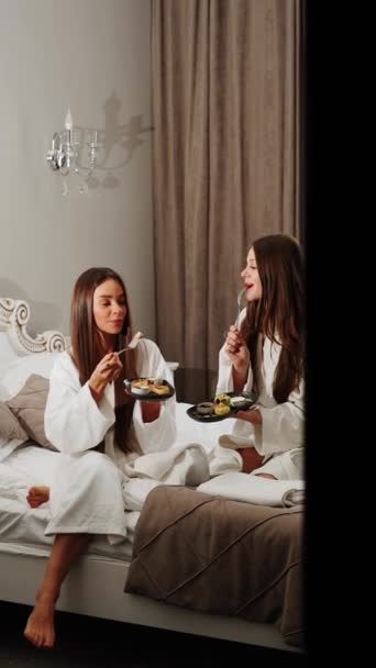 Bornozlu iki genç kadın yatakta kahvaltı yapıyor. Sağlıklı kahvaltı peynirli kekler. Otel tatili konsepti. Dikey video - Video, Çekim