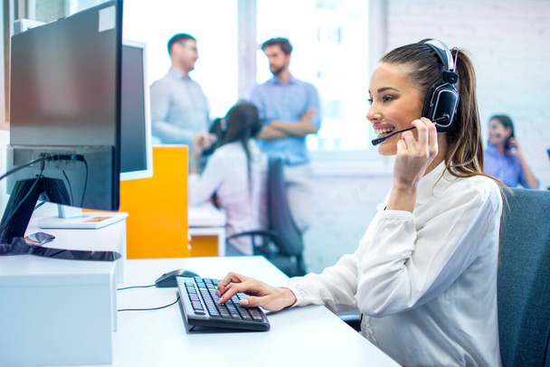 Sonriente mujer operador de línea directa con auriculares utilizando el ordenador en la oficina. - Foto, Imagen