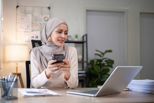 Feliz mujer de negocios musulmana usando hijab en el trabajo Utilice su computadora portátil y teléfono inteligente para trabajar en finanzas y marketing en la oficina.. - Foto, imagen