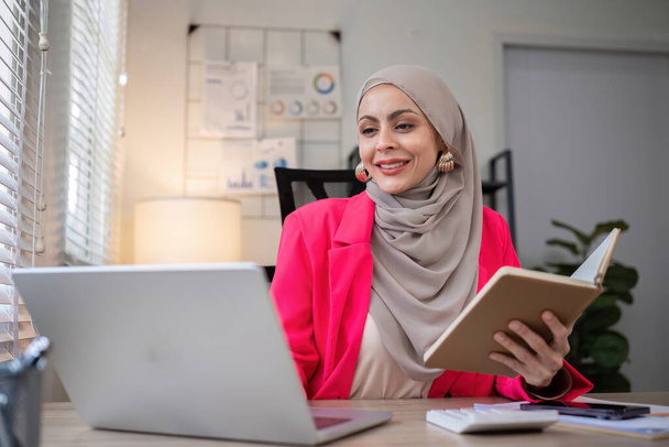 Молода мусульманська бізнес-леді робить нотатки про бізнес-репортажі і працює на ноутбуці за столом в офісі. - Фото, зображення