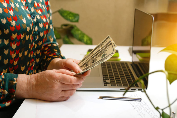 Рука женщины считает доллары, работая над ноутбуком в яркой офисной обстановке, символизируя финансовый менеджмент и инвестиционные решения. - Фото, изображение