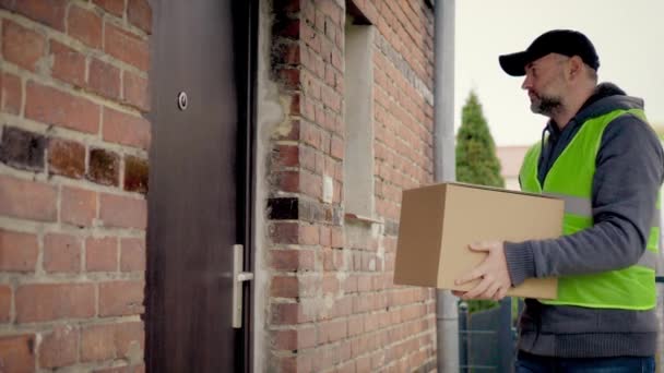 Young woman opens doors of her house and meets delivery man who gives her cardboard box postal package and writes signature in document. Concept of courier, home delivery, e-commerce shipping - Footage, Video