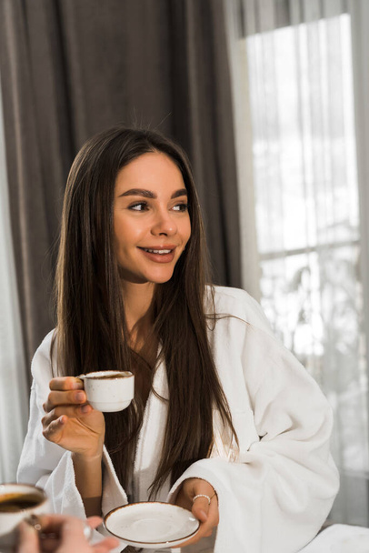 Young women in bathrobes have breakfast in bed, drink coffee and fool around. Breakfast cheesecakes and fruits with coffee. Hotel holiday concept - Photo, Image