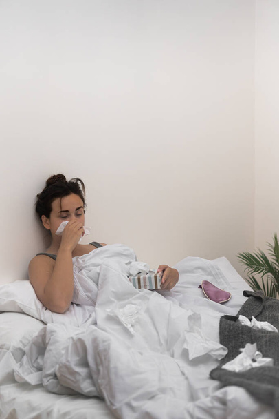 A letto, una giovane donna malata si pulisce il naso con dei tessuti, tenendo delle gocce mediche per alleviare la malattia..  - Foto, immagini