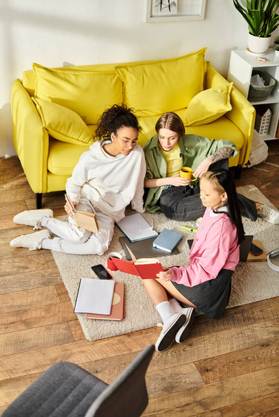 Drei junge Mädchen verschiedener Rassen, die auf dem Boden sitzen und in Bücher vertieft sind, lernen gemeinsam zu Hause in einem Zeichen der Freundschaft und des Engagements für die Bildung. - Foto, Bild