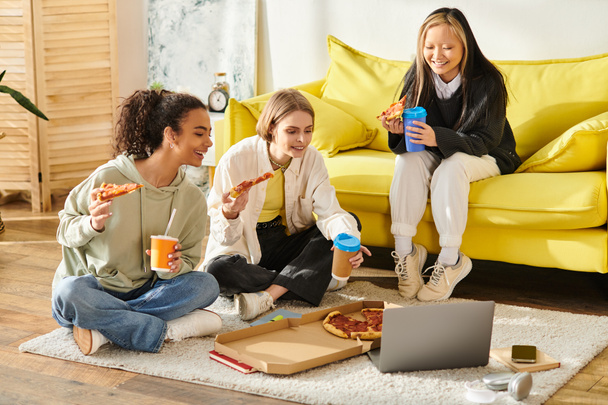 Τρία έφηβα κορίτσια διαφορετικών φυλών κάθονται στο πάτωμα, απολαμβάνοντας πίτσα και καφέ μαζί σε ένα άνετο περιβάλλον. - Φωτογραφία, εικόνα