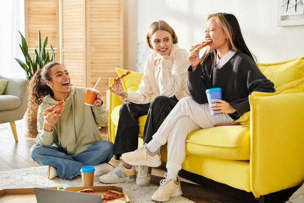 3人の多様な若い女性が居心地の良いソファーでピザとコーヒーを楽しみ,食べ物と笑いを結びつけています.. - 写真・画像
