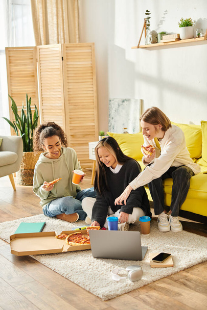 Adolescentes de diferentes razas se sientan juntas en el suelo, riendo y comiendo pizza, disfrutando de un momento de amistad. - Foto, Imagen
