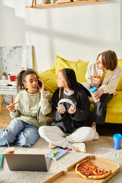Trzy zaangażowane nastolatki z różnych środowisk etnicznych cieszą się zwykłą pizzą siedząc na podłodze. - Zdjęcie, obraz