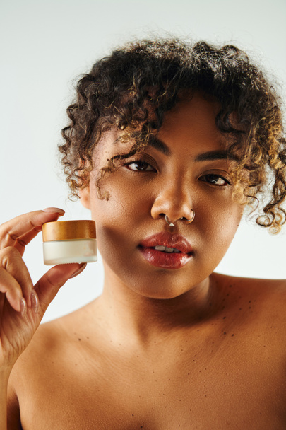 Una hermosa mujer afroamericana sostiene un frasco de crema frente a su cara. - Foto, Imagen