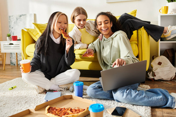 Τρία έφηβα κορίτσια διαφορετικών εθνικοτήτων κάθονται στο πάτωμα, απολαμβάνοντας την πίτσα και χρησιμοποιώντας ένα φορητό υπολογιστή μαζί. - Φωτογραφία, εικόνα