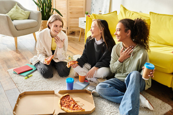multiculturele tienermeisjes zitten op de vloer, genieten van pizza samen in een gezellige omgeving. - Foto, afbeelding