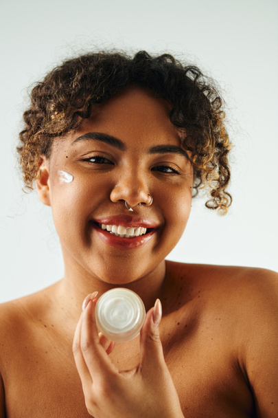 Afro-Amerikaanse vrouw energiek houdt crème fles tegen levendige achtergrond. - Foto, afbeelding