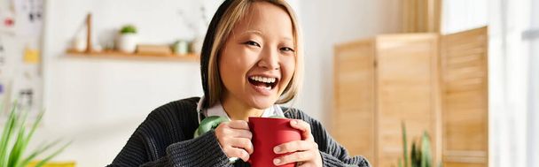 Uma jovem, desfrutando de um momento de riso, segura uma xícara vermelha na mão enquanto estuda em casa - Foto, Imagem