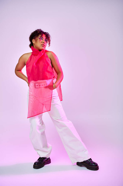 An African American woman poses confidently in a pink shirt and white pants against a vibrant backdrop. - Φωτογραφία, εικόνα