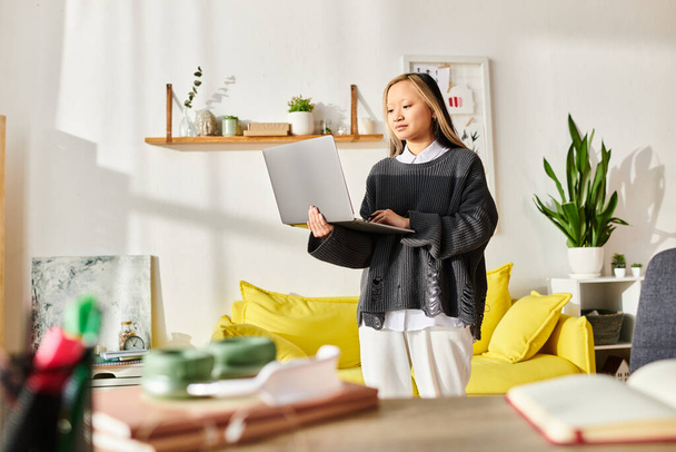Eine junge Asiatin steht elegant in einem modernen Wohnzimmer und ist in E-Learning vertieft, während sie einen Laptop in der Hand hält.. - Foto, Bild